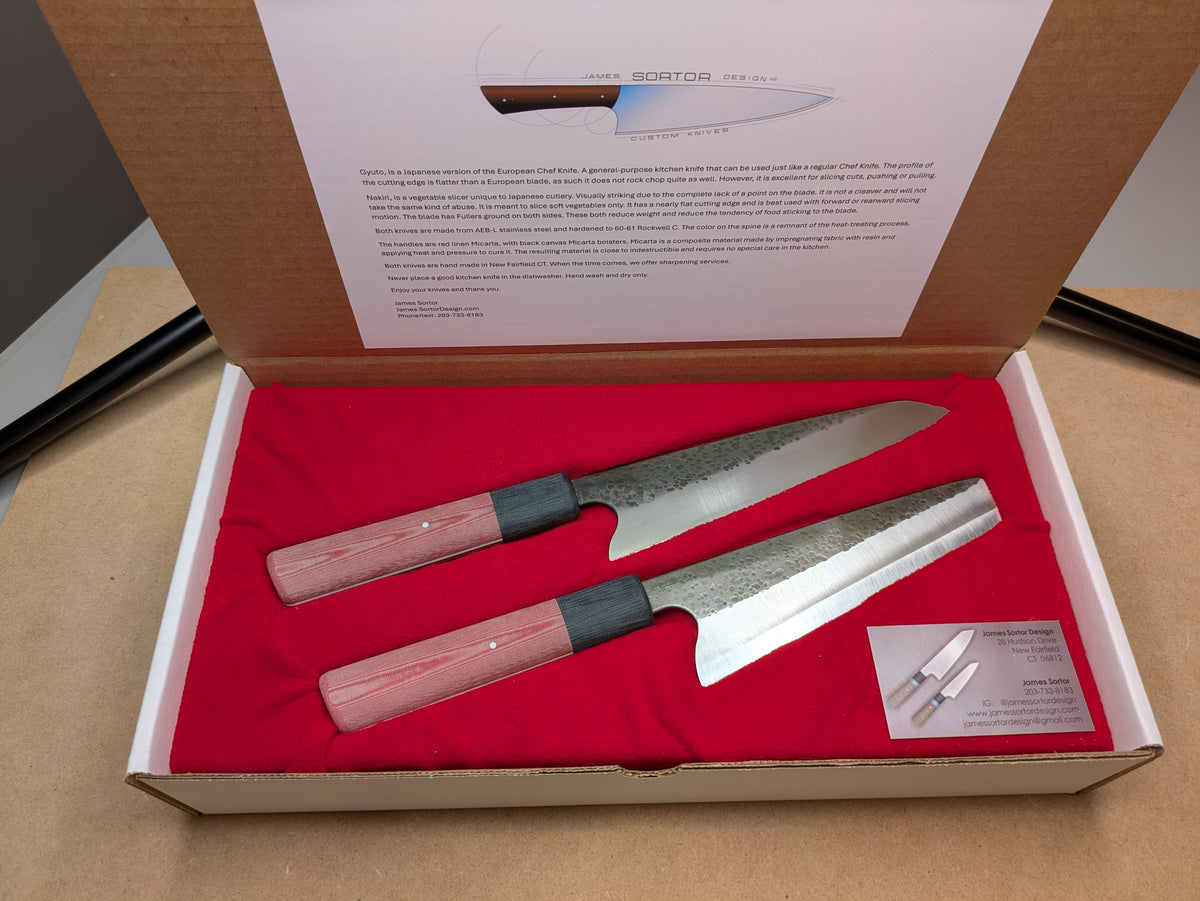 Two Japanese kitchen knives with patterned steel blades and red linen Micarta handles, presented in an open white gift box with a red fabric lining and an interior product card.