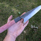 A hand holding a Japanese-style fixed blade knife outdoors on a grassy lawn. The knife features a satin-finished steel blade with a dark etched pattern near the base, a black bolster, and a red linen Micarta handle.