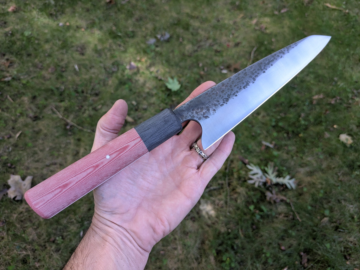A hand holding a Japanese-style fixed blade knife outdoors on a grassy lawn. The knife features a satin-finished steel blade with a dark etched pattern near the base, a black bolster, and a red linen Micarta handle.