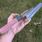 A hand holding a Japanese-style kitchen knife with a red linen Micarta handle and a stainless-steel blade, set against a background of green grass.