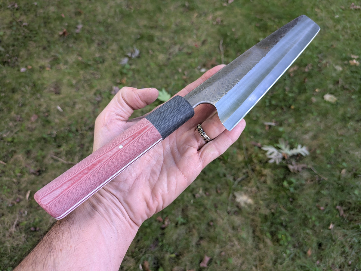 A hand holding a Japanese-style kitchen knife with a red linen Micarta handle and a stainless-steel blade, set against a background of green grass.