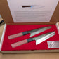 Two Japanese kitchen knives with patterned steel blades and red linen Micarta handles, presented in an open white gift box with a red fabric lining and an interior product card.