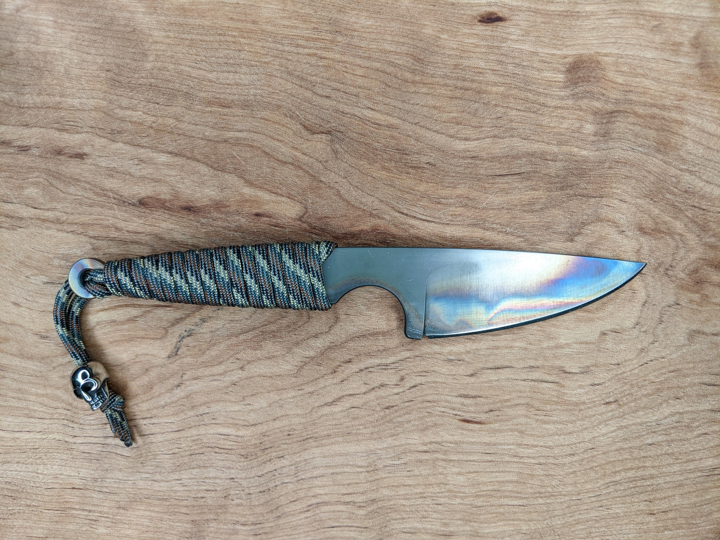 small knife with heat-treating colors and paracord wrapped handle with a metal skull bead, on wooden background